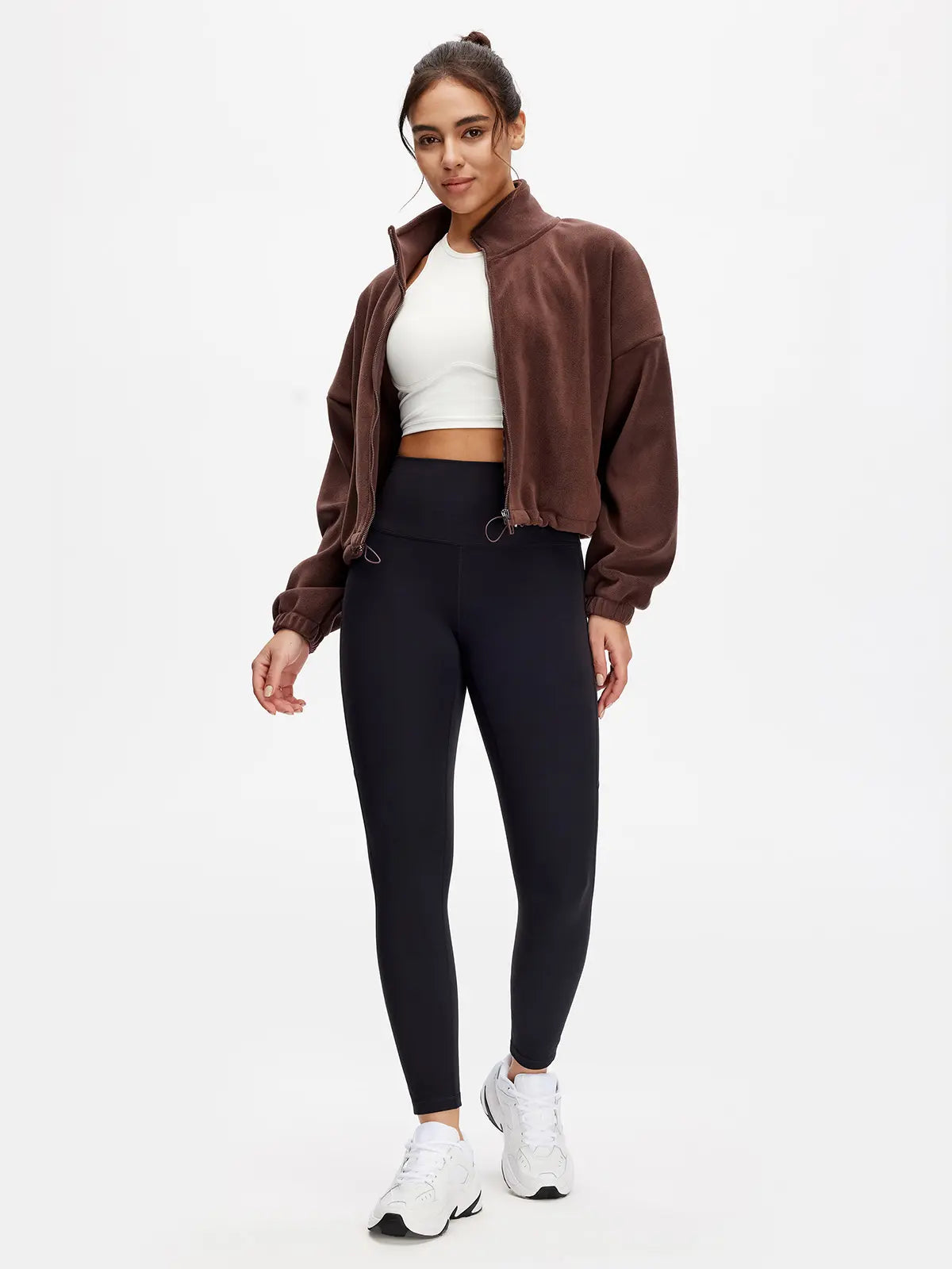 Woman wearing a brown jacket, white top, black leggings, and white sneakers on a white background